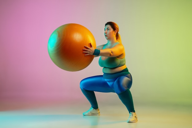 Entrenamiento del modelo femenino caucásico joven del tamaño extra grande en la pared verde púrpura del gradiente en luz de neón. Haciendo ejercicios de entrenamiento con fitball.