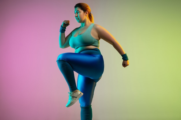 Entrenamiento del modelo femenino caucásico joven del tamaño extra grande en la pared verde púrpura del gradiente en neón. Haciendo ejercicios de estiramiento. Concepto de deporte, estilo de vida saludable, cuerpo positivo, igualdad.