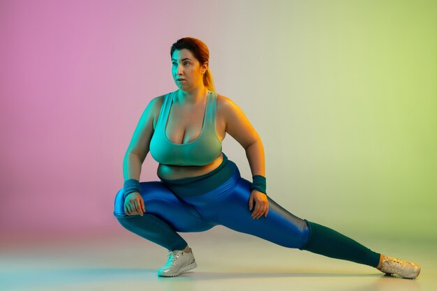 Entrenamiento del modelo femenino caucásico joven del tamaño extra grande en la pared verde púrpura del gradiente en neón. Haciendo ejercicios de estiramiento. Concepto de deporte, estilo de vida saludable, cuerpo positivo, igualdad.