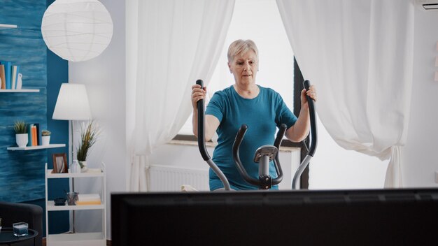 Entrenamiento de personas mayores en bicicleta estacionaria electrónica para hacer ejercicio físico y actividad. Mujer mayor que usa una máquina de ciclismo cardiovascular para entrenar los músculos de las piernas con gimnasia en casa.