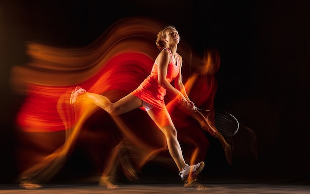 Entrenamiento de tenista profesional aislado sobre fondo negro de estudio en luz mixta. Mujer en traje deportivo practicando.