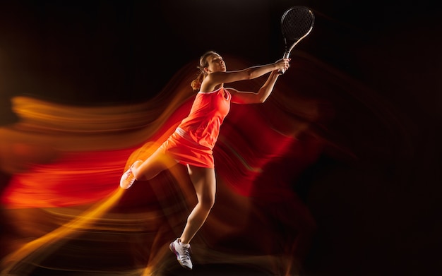 Entrenamiento de tenista profesional aislado sobre fondo negro de estudio en luz mixta. Mujer en traje deportivo practicando.