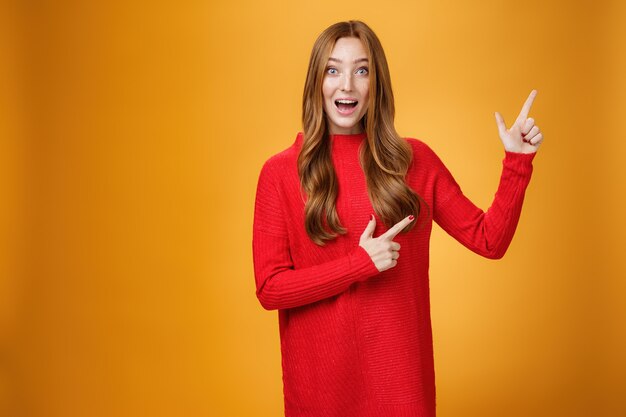 Entusiasta y ambiciosa mujer pelirroja linda con pecas en vestido rojo apuntando a la esquina superior derecha sonriendo asombrada y curiosa mientras explora un nuevo espacio impresionante posando sobre fondo naranja