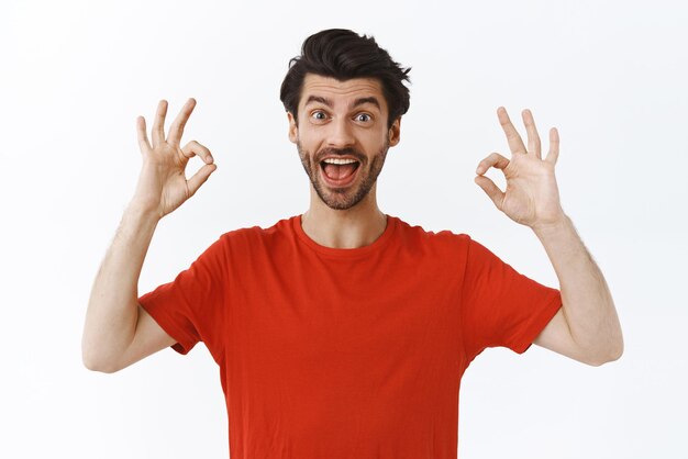 Entusiasta apuesto hombre barbudo con camiseta roja que muestra el signo bien, está bien o está bien y sonríe complacido dar aprobación recomendar producto increíble dar retroalimentación positiva fondo blanco