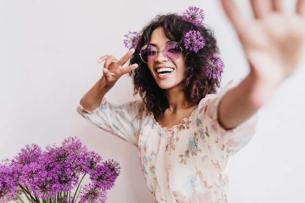 Entusiasta chica africana en elegante vestido haciendo selfie con alliums. Espectacular joven negra divirtiéndose durante la sesión de fotos en interiores con flores de color púrpura.