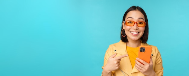 Entusiasta chica asiática con elegantes gafas de sol apuntando con el dedo al teléfono inteligente que muestra el teléfono móvil de pie sobre fondo azul.