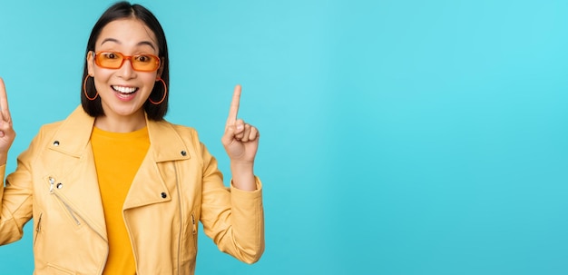 Entusiasta chica asiática con gafas de sol apunta con el dedo hacia arriba muestra un cartel o un logotipo en los soportes superiores sobre fondo azul Copiar espacio