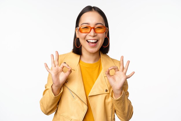 Entusiasta chica asiática con gafas de sol muestra bien ok firmar en aprobación sonriendo y riendo aprobar recomendar algo elogio y cumplido de pie sobre fondo blanco