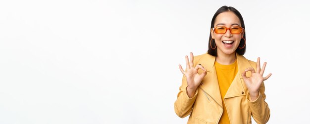 Entusiasta chica asiática con gafas de sol muestra bien ok firmar en aprobación sonriendo y riendo aprobar recomendar algo elogio y cumplido de pie sobre fondo blanco