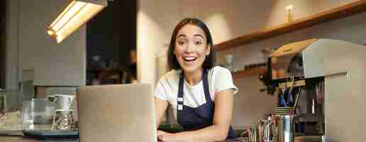 Foto gratuita entusiasta chica asiática en uniforme de cafetería barista trabajador con portátil mirando feliz y sorprendido en