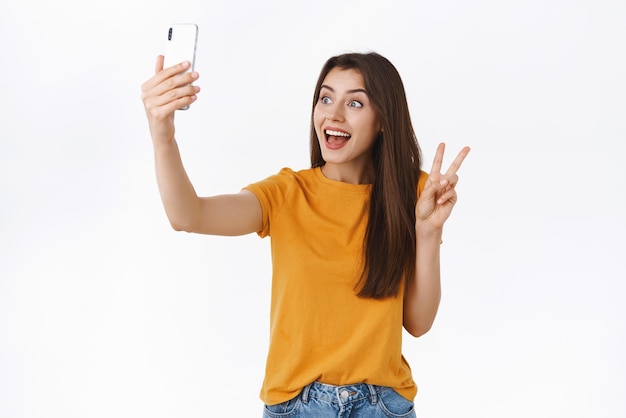 Entusiasta chica bastante tonta en camiseta amarilla levantando el teléfono móvil tomando selfie en el teléfono inteligente que muestra el signo de la paz o la victoria mira la cámara del teléfono divertida de pie fondo blanco