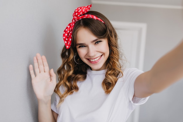 Entusiasta chica blanca con cabello ondulado brillante tomando una foto de sí misma en casa. Foto interior de modelo femenino feliz con cinta roja haciendo selfie.