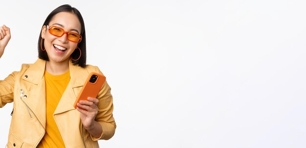 Entusiasta chica coreana con gafas de sol sosteniendo un teléfono inteligente celebrando y bailando riendo feliz con el teléfono móvil de pie sobre fondo blanco.