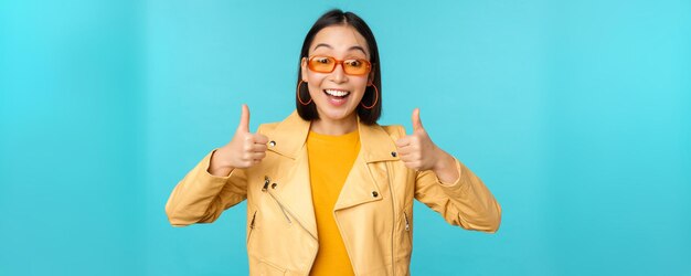 La entusiasta chica coreana se ve emocionada y muestra el pulgar hacia arriba aprueba algo impresionante, excelente elección y calidad sobre fondo azul.