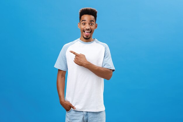 Entusiasta, complacido y encantado chico de piel oscura con barba y peinado afro de pie alegre sobre fondo azul sonriendo alegremente apuntando a la esquina superior izquierda sosteniendo la mano en el bolsillo.