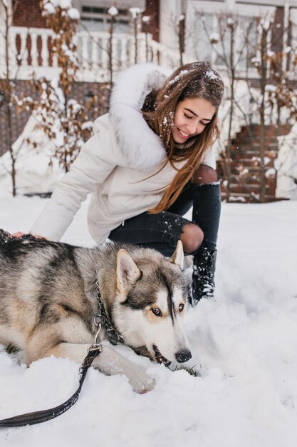 Entusiasta dama europea mira con amor a su perro, cansada después del juego en la nieve. Chica guapa en elegante atuendo blanco jugando con husky en día helado.