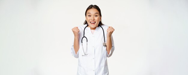 Entusiasta doctora asiática celebrando luciendo esperanzada y feliz ante la cámara triunfando de pie en uniforme médico contra fondo blanco