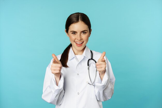 Entusiasta doctora sonriendo señalando con el dedo a la cámara felicitando invitando a los pacientes a c...
