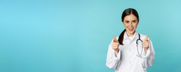 Foto gratuita entusiasta doctora sonriendo señalando con el dedo a la cámara felicitando invitando a los pacientes a cli