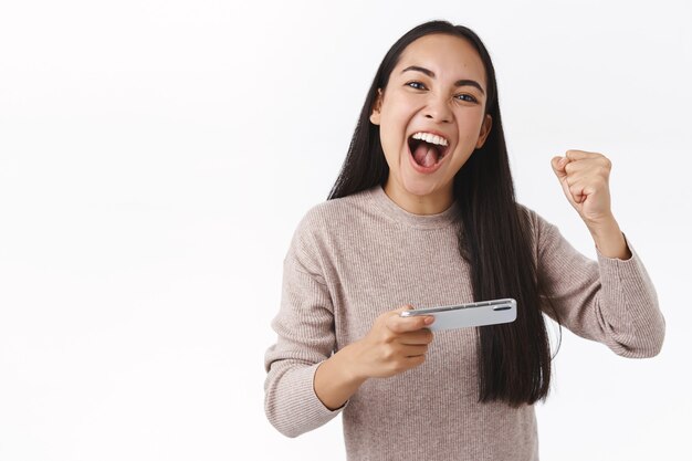 Entusiasta, guapa, alegre, joven asiática, ganar el juego, batir la puntuación, golpear el puño triunfando y gritando que sí mientras se anima a sí misma, sosteniendo el teléfono inteligente horizontalmente, terminando el nivel difícil