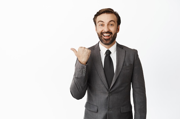 Entusiasta hombre de negocios barbudo señalando con el dedo a la izquierda y un hombre corporativo sonriente mostrando un anuncio de pie en traje sobre fondo blanco.