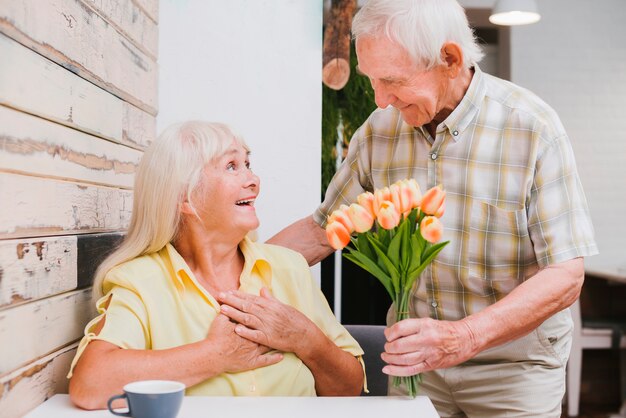 Entusiasta hombre senior presentando flores a la mujer en la cafetería