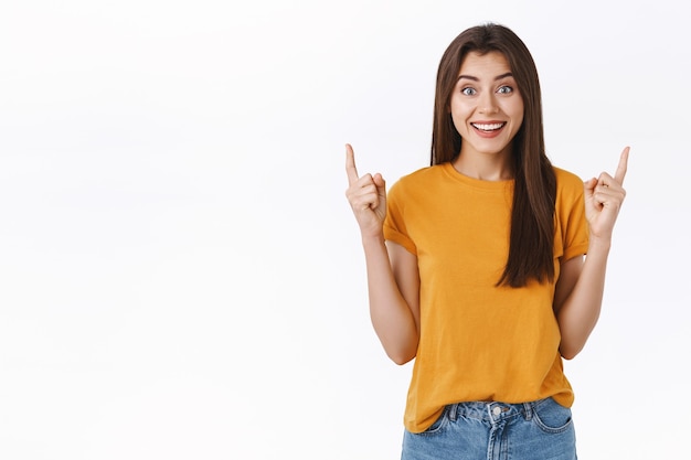 Entusiasta, impresionada y emocionada sonriendo, feliz mujer morena con camiseta amarilla, riendo y sonriendo emocionada, no puedo esperar para probar una increíble propuesta de venta, señalando un anuncio genial