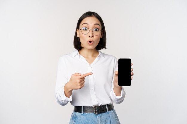 Entusiasta joven asiática señalando con el dedo a la pantalla del teléfono inteligente que muestra publicidad en el fondo blanco del teléfono móvil