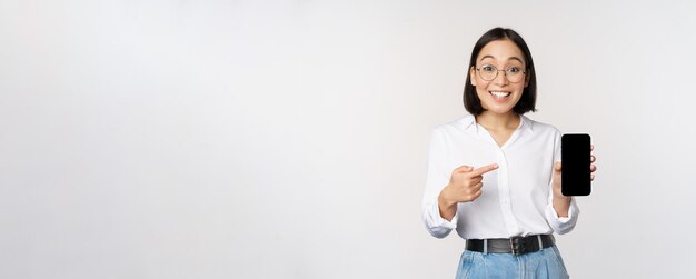 Entusiasta joven asiática señalando con el dedo a la pantalla del teléfono inteligente que muestra publicidad en el móvil