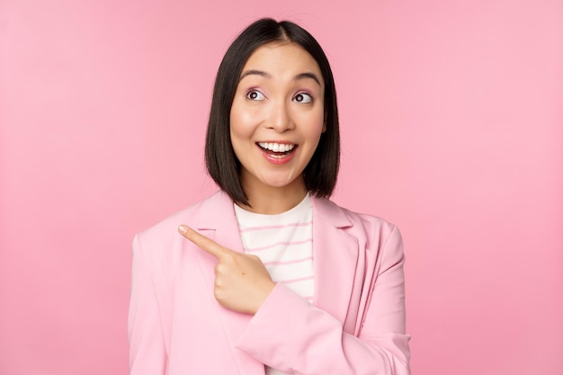 Entusiasta joven empresaria asiática empleada de oficina señalando con el dedo a la izquierda mirando el anuncio de pancarta con una sonrisa feliz que muestra el fondo rosa del anuncio