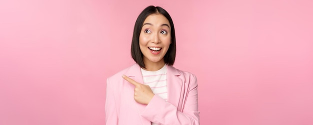 Entusiasta joven empresaria asiática empleada de oficina señalando con el dedo a la izquierda mirando el anuncio de pancarta con una sonrisa feliz que muestra el fondo rosa del anuncio