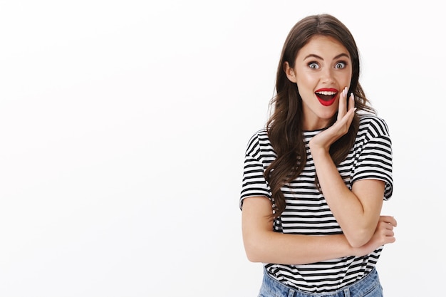 Entusiasta mujer alegre y divertida escuchar chismes, sonriendo entretenido y alegre, jadeo sorprendido, mirar la cámara alegre, reaccionar fascinado descubrir maravillosa promoción impresionante, pared blanca