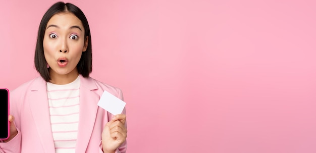 Entusiasta mujer de negocios asiática que muestra la pantalla del teléfono móvil y la tarjeta de crédito mirando asombrada a la cámara de pie sobre fondo rosa