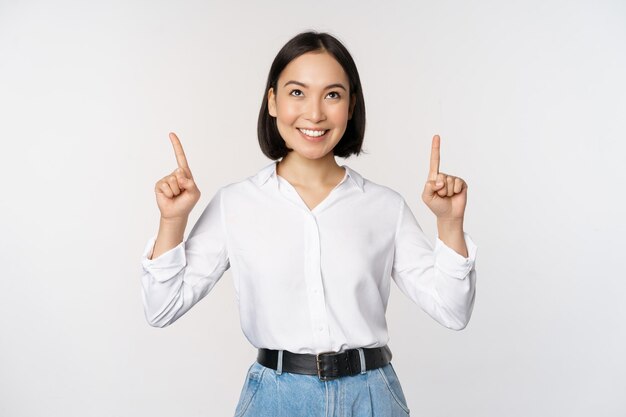 Entusiasta mujer de negocios asiática señalando mirando hacia arriba con una cara sonriente feliz que muestra el logotipo de la empresa o una pancarta de pie sobre fondo blanco