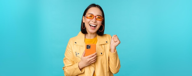 Entusiasta niña asiática sonriente con gafas de sol sosteniendo un teléfono móvil y bailando riendo con un teléfono inteligente de pie sobre fondo azul.