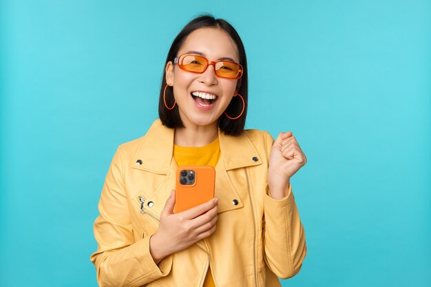 Entusiasta niña asiática sonriente con gafas de sol sosteniendo un teléfono móvil y bailando riendo con un teléfono inteligente de pie sobre fondo azul.