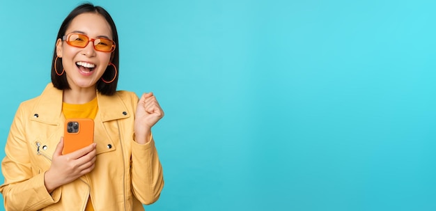 Entusiasta niña asiática sonriente con gafas de sol sosteniendo un teléfono móvil y bailando riendo con un teléfono inteligente de pie sobre fondo azul.