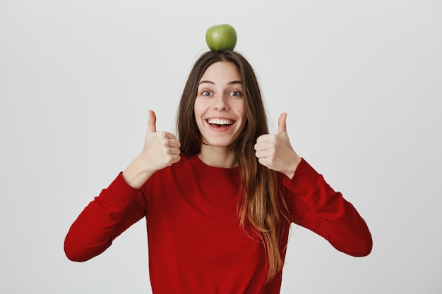 Foto gratuita entusiasta niña sonriente con manzana verde en la cabeza mostrando el pulgar hacia arriba