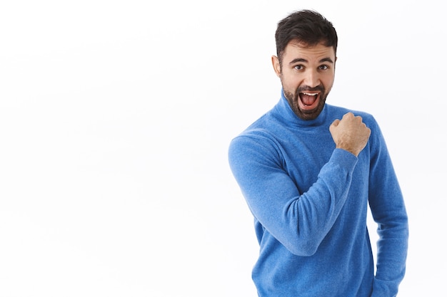 Entusiasta y optimista, feliz y confiado chico caucásico alentando a seguir adelante, puñetazos y sonriendo, sentirse optimista listo para ir hacia el sueño, vestido para el éxito, de pie en la pared blanca