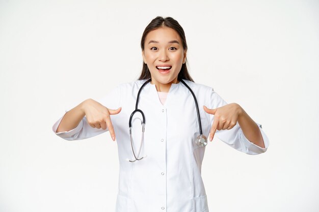 Entusiasta personal médico, trabajadora sanitaria asiática, apuntando con el dedo hacia abajo y sonriendo asombrado, mostrando descuentos, venta en clínica, fondo blanco.