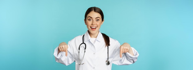 Entusiasta trabajadora de la salud joven doctora en bata blanca apuntando con el dedo hacia abajo y sonriendo sh