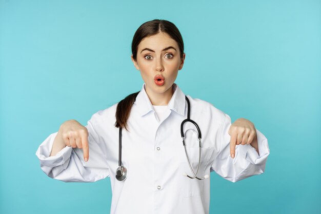 Entusiasta trabajadora de la salud joven doctora en bata blanca apuntando con el dedo hacia abajo y sonriendo...