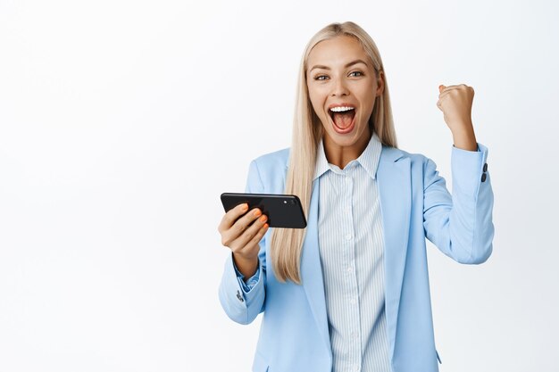 Entusiasta vendedora animando usando el teléfono móvil y celebrando lograr la meta en el teléfono inteligente de pie en traje sobre fondo blanco.