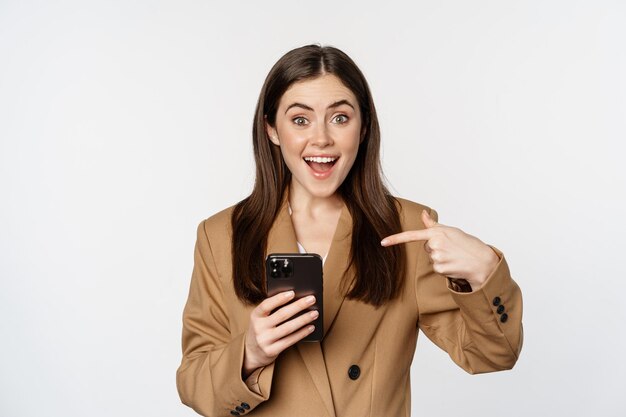 Entusiasta vendedora mujer de negocios señalando con el dedo el teléfono móvil y sonriendo mostrando en celular...