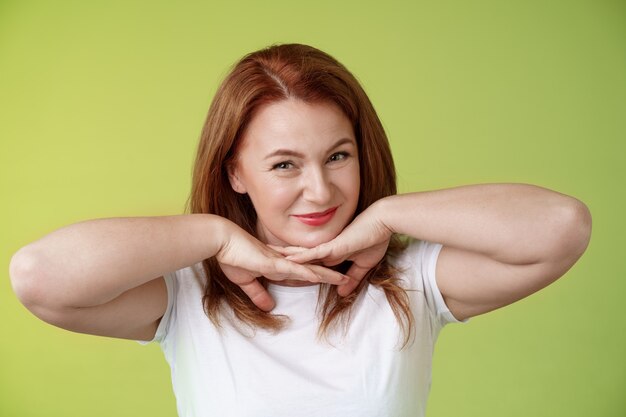 Envejecimiento cosmetología concepto de bienestar feliz mujer pelirroja segura de sí misma tomarse de las manos debajo de la línea de la mandíbula sonriendo mostrando arrugas faciales imperfectas aplicando productos para el cuidado de la piel pared verde