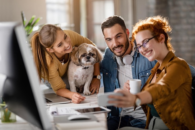 Equipo de alegres colegas de negocios con un perro divirtiéndose mientras toman selfie en la oficina