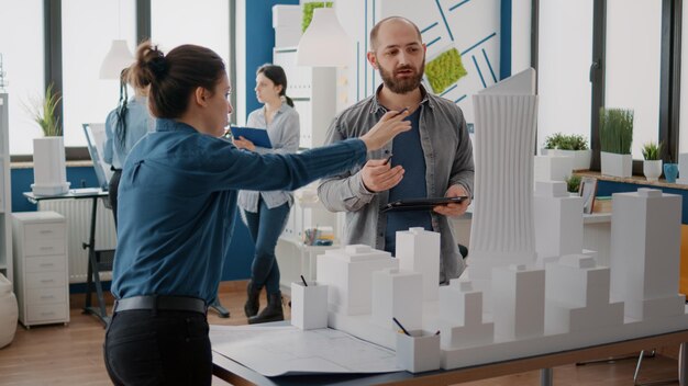 Equipo de compañeros de trabajo analizando maqueta y modelo de construcción para planificar la construcción en tableta. Personas arquitectónicas trabajando juntas, diseñando estrategia urbana y desarrollo inmobiliario.