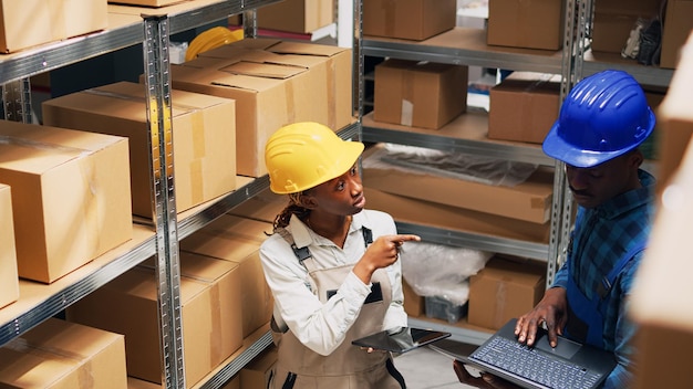 Equipo de empleados que verifican existencias en computadoras portátiles y tabletas, utilizando dispositivos para planificar la logística de mercancías en el área del almacén. Jóvenes trabajando con envases de cartón y productos de carga. Disparo de trípode.