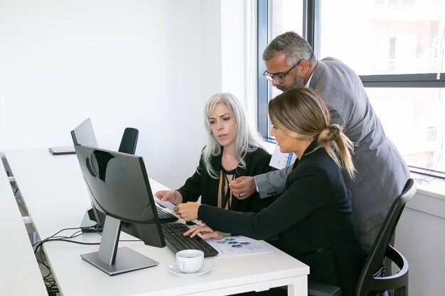 Equipo empresarial de tres informes de análisis, sentado en el lugar de trabajo con monitores juntos, sosteniendo, revisando y discutiendo documentos con gráficos. Copie el espacio. Concepto de lugar de trabajo inclusivo