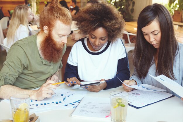 Equipo de expertos en marketing que desarrollan la estrategia empresarial en el café Mujer africana que presenta un plan de negocios a su socio con barba roja en tableta digital mientras su colega asiático analiza gráficos